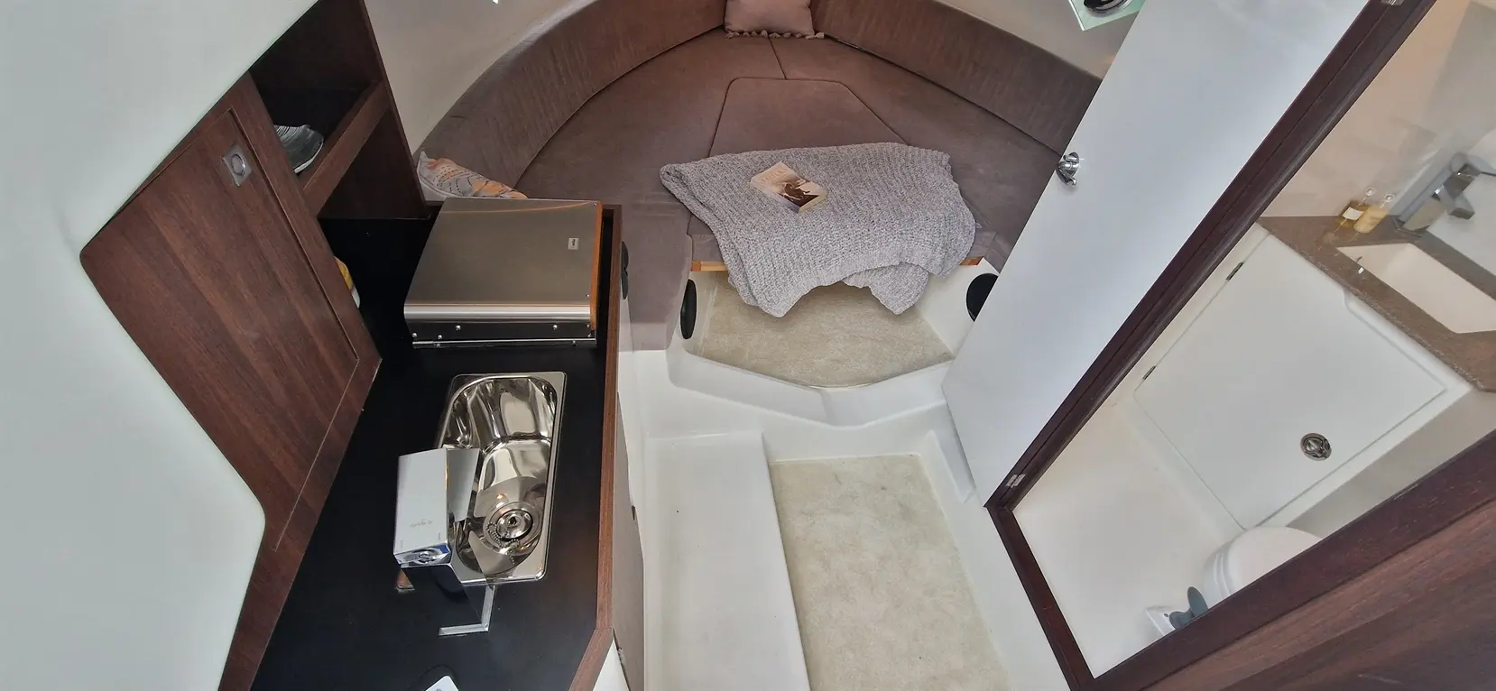 Interior of a yacht showcasing a compact kitchen sink, grey blanket, book, and a seating area with a padded berth.